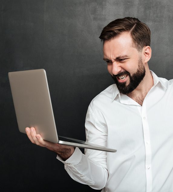 portrait-agressive-bearded-man-holding-silver-personal-computer-throwing-punch-screen-isolated-dark-gray-wall